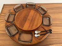 Top view of teak Lazy Susan with seven smoky grey glass dishes arranged around it and two small serving spoons placed in the empty slot.