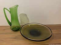 Both green glass bowl and matching tall pitcher displayed side by side on a wooden surface against a white background, clear view of shape and color.