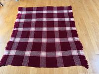 Full view of the wool blanket on a wood floor showing cranberry base with pink and cream plaid pattern and fringed edges.