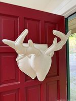 White felt deer head wall/door decoration photographed hanging on a red door, showing front and side views with visible antlers and shield plaque mounting.