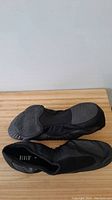 Pair of black Bloch Flex Studio fitness slippers on a wooden surface, bottom and side views showing sole and design.