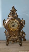 Front view of ornate brass mantle clock showing detailed floral scrollwork and clock face with glass cover.