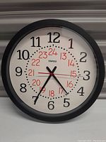 Front view of Staples wall clock showing black frame, white face, black large hour numbers and red 24-hour numbers.