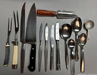 Full assortment of flatware, kitchen knives, forks, spoons, ladles and vegetable peeler laid out in a single row.