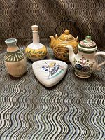 Five ceramic items arranged on patterned fabric, includes the wall pocket, two teapots, cruet, and vase.