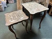 Pair of two nesting tables with cream toned faded decorative patterned tops and bronze tone curved slender legs shown upright on carpet.