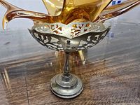 Front and side view of the silverware footed compote dish showing ornate cut-out floral pattern and round base, placed on wood surface near amber glass object.