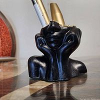 Front view of black makeup holder shaped like a girl's head resting on her hands, holding three makeup items.