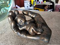 Bronze effect baby sculpture lying in a sculpted hand, showing detail of baby and hand, placed on a table surface.