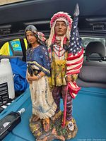 Front view of vintage figurine showing Native American chief in feathered headdress holding American flag with native woman beside him.