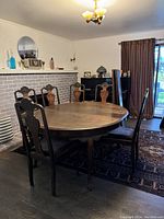 Photo of round antique dining table with five extension leaves, surrounded by six chairs varying with and without arms, positioned in a dining room with a rug underneath and a decorative fireplace in the background.