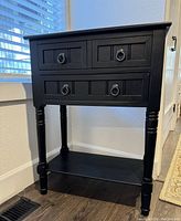 Front and side view of the black wooden end table showing all four turned legs and three drawers with metal ring pulls, plus bottom shelf.