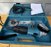 Open Makita carrying case showing angle grinder, cutting wheel, side handle, wrench, guard, and manual.