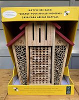 Front view of the native bee barn showing natural bamboo tubes for bee nesting and wooden block with drilled holes, housed in a wood structure with red roof accents and engraved bees.