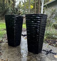 Two tall black glazed ribbed planters on a concrete surface outdoors, small black plastic pedestal stand pieces beside them.