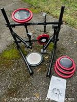 Photo showing Remo practice drum kit with 5 pieces mounted on a black frame outdoors. Includes four red edged drum practice pads and one white mesh kick drum pad. Instruction sheet and adjustment tool visible on ground.