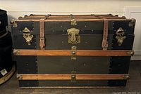 Front view showing black metal panels, three horizontal wood slats, central brass lock, two fasteners for leather straps and brass corner protectors.