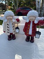 Two resin children figurines side by side, one in white coat and the other in red coat, both holding small lanterns.