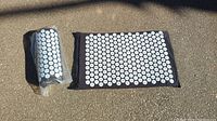 Overall view of the acupressure pillow and mat set outdoors, showing the black base with multiple white spiked plastic discs on surface. The mat is laid flat and pillow is rolled in plastic.