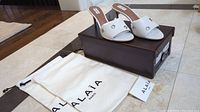 White Alaia high heel sandals on top of brown box with white Alaia dust bag beside it.