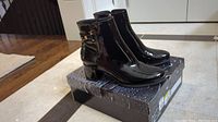 Pair of glossy black Artica waterproof ankle boots on shoebox, showing front and side views with decorative buckle and zipper details.