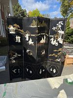 Full view of four-panel black lacquer decorative screen with gold painted landscape, coral and white inlay figures and motifs, standing outdoors in sunlight.