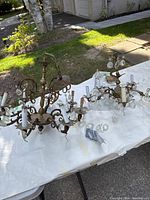 Pair of vintage ornate bronze chandeliers with multiple arms and candle-style bulb sockets, resting on a white table outdoors. Crystals visible but some missing.