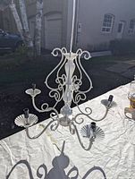 White chandelier with metal scrollwork and clear crystal center photographed outdoors on white sheet, showing five arm holders each with decorative bobeches.