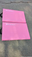 Two folded bright pink exercise mats placed on asphalt surface showing black handles and white branding.