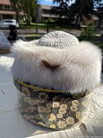 Front view of the fur rimmed hat with crocheted top inside the protective box with daisy print, showing the hat's wide fur rim.