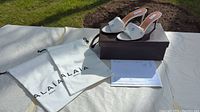 Pair of white Alaia high heel sandals on dark brown box with two white dust travel bags and paperwork on a white cloth outdoors on grass background.