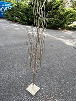 Full front view of free standing gold metal tree showing its height and thin branch extensions on a square base placed on pavement outside.