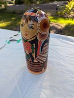 Front and angled views of a single wooden Russian nesting doll on a white surface outdoors showing detailed hand-painted facial features and traditional costume with floral and geometric designs.