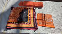 Three scarves laid out on a white cloth with sunlight showing colors and textures. One folded orange textured polyester scarf, one small square silk scarf, and one large folded silk scarf with multicolored border.