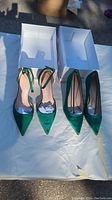 Both pairs of emerald green satin heels side by side in their white shoeboxes outdoors on a white surface.