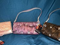 Three clutch purses laid out against blue fabric background in a single photo, showing their colors and designs clearly.