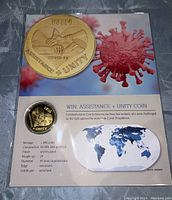 Top-down photo of commemorative 24kt gold foil coin with design of clasped hands, text 'WIN ASSISTANCE + UNITY', and Covid-19 virus imagery on certificate background.
