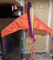 Orange airplane-shaped kite with purple edges and yellow vertical tail section, displayed indoors suspended against a door.