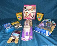 Photo showing all items in the lot arranged on a blue fabric background including the Do Dats DIY kit, crayons, baby doll clothes, baby nasal care, thumb sleeves, and door stoppers.