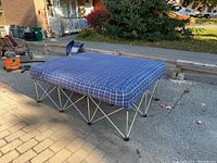 Photo of the queen size air bed fully inflated and set up outdoors on a paved surface. The blue plaid cover and folding metal frame are clearly visible.