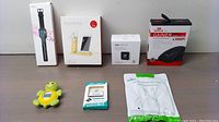 All main items from the lot displayed on a table: smartwatch, smartphone stand, air quality monitor, gaming mouse, turtle device, adapter, and cable pack.