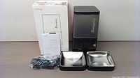 Automatic cat feeder unit with translucent black container, FEELNEEDY branded, two square stainless steel bowls with black bases, power adapter, and user manual.