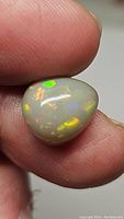 Close-up view of the pear-shaped semi-black Ethiopian opal held between thumb and finger showing multicolored flashes of green, yellow, orange, and red.