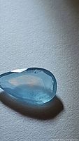 Close-up photo of a pear-shaped light blue aquamarine gemstone with translucent clarity and visible natural inclusions, placed on a textured surface.