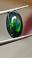 Close-up view of the 1.11ct natural black opal oval cabochon gemstone with visible multicolored play-of-color held in tweezers.