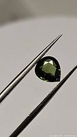 Close-up of pear-shaped dark green tourmaline held by tweezers showing faceted cuts and clarity.