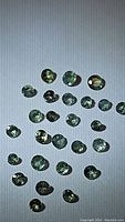 Multiple small round faceted greenish to light green-yellow Alexandrite gemstones on white background, showing variations in size and slight color change properties.