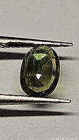 Close-up of 0.77 carat oval cut green sapphire held with tweezers showing color and clarity.