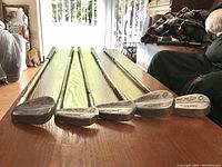 Five vintage stainless steel golf clubs arranged on a wooden floor showing club heads and shafts.
