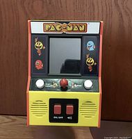 Front view of handheld Pac-Man game, powered off, showing control buttons, joystick, and yellow and orange arcade-style casing with Pac-Man characters.
